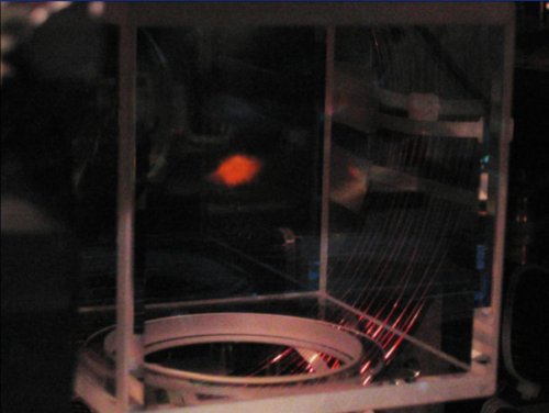  Fig. 2. Photographie du piège magnéto-optique dans lequel sont confinés les atomes froids. On aperçoit en bas à droite une partie des enroulements conducteurs créant les champs magnétiques qui concourent au confinement des atomes. La tache rouge est due à la diffusion de la lumière du laser de pompage optique par la mélasse d’atomes froids. Crédit Robin Kaiser. 